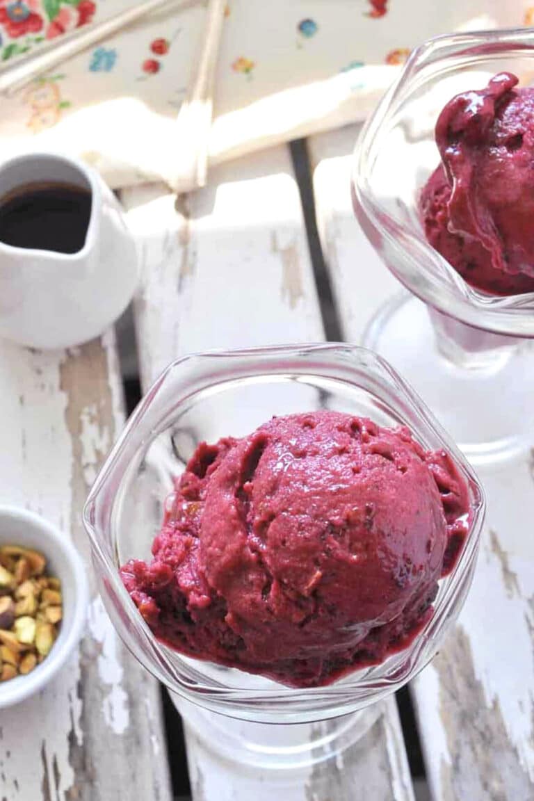 Two glass dishes filled with scoops of deep purple berry sorbet and Quick Cherry Ice Cream sit on a white wooden surface. A small bowl of pistachios and a pitcher of dark syrup nearby glisten in the bright natural light.