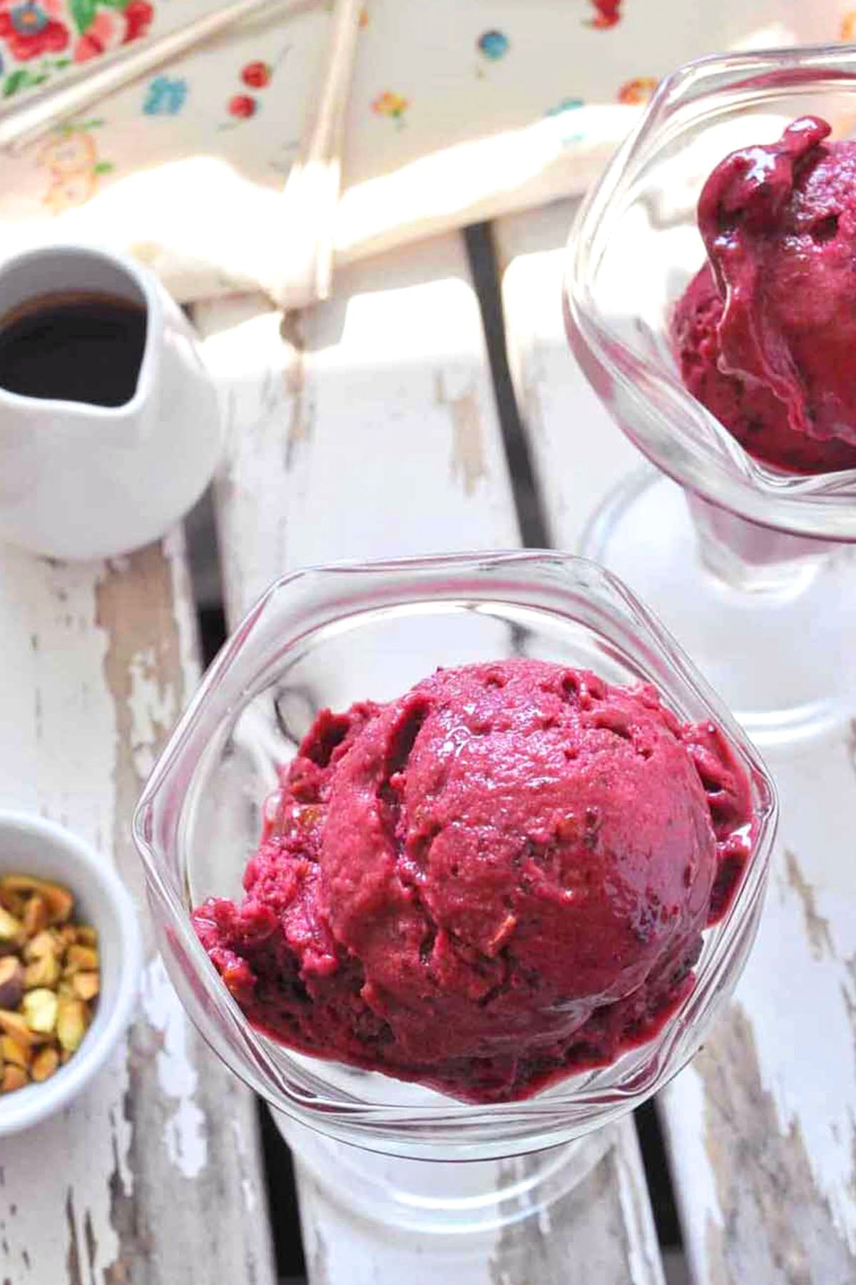 Two glass dishes filled with scoops of deep purple berry sorbet and Quick Cherry Ice Cream sit on a white wooden surface. A small bowl of pistachios and a pitcher of dark syrup nearby glisten in the bright natural light.