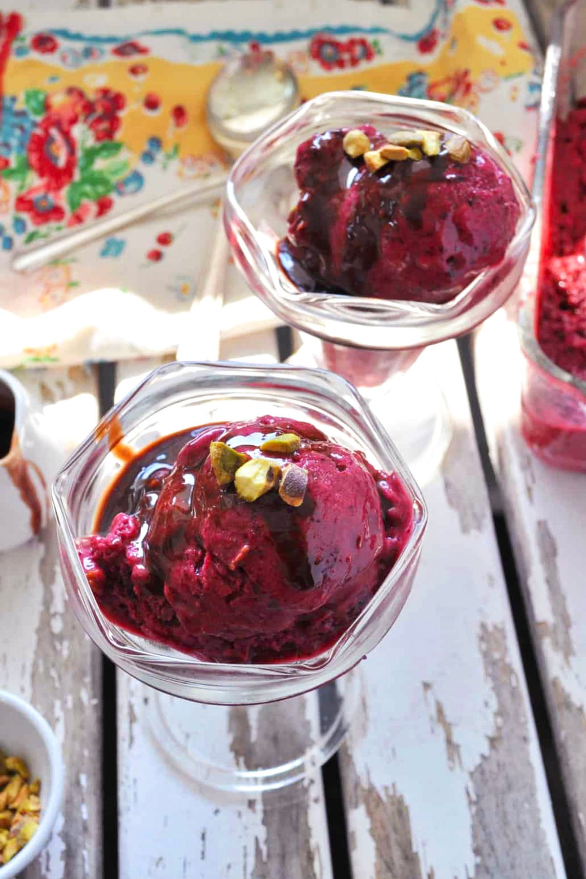 Two glass bowls with scoops of dark purple quick cherry ice cream, drizzled with chocolate sauce and topped with chopped pistachios, are on a white wooden table with a floral napkin nearby.