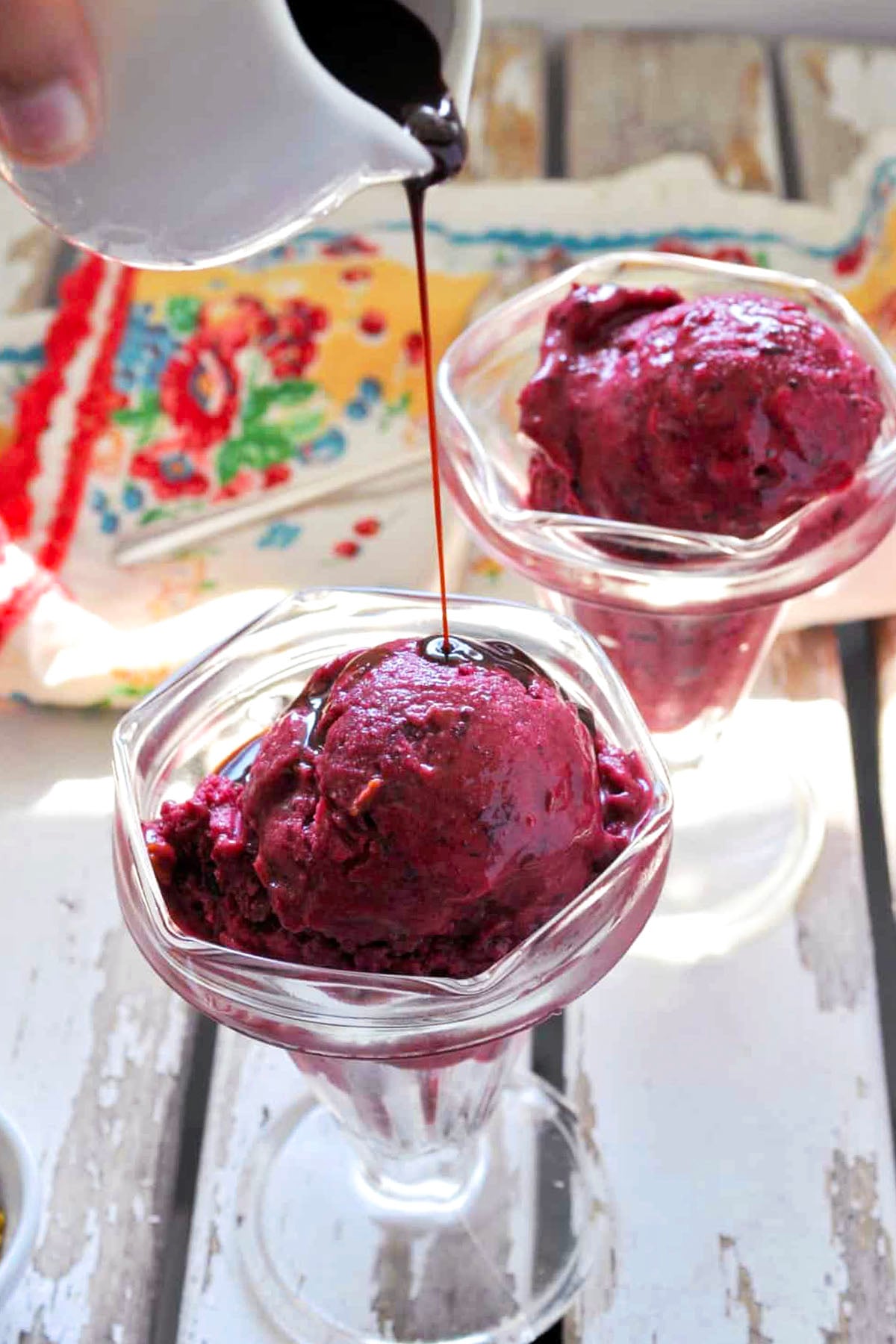 Two glass dessert cups filled with rich Quick Cherry Ice Cream are topped as chocolate sauce is poured from a small white pitcher. A colorful cloth brightens the background.