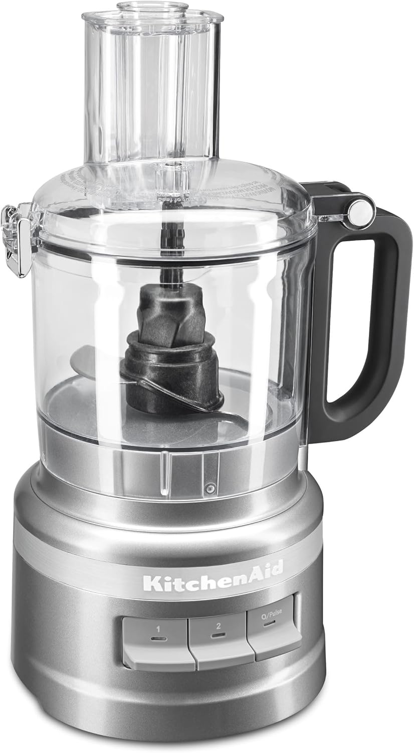 A silver KitchenAid food processor with a clear plastic bowl, black handle, and three control buttons on the base—perfect for making homemade cherry vanilla ice cream—is shown on a white background.