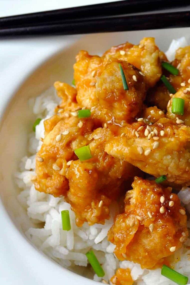 A close-up of crispy oven-fried orange chicken pieces topped with sesame seeds and chopped green onions, served over a bed of white rice, with black chopsticks resting on the plate edge.