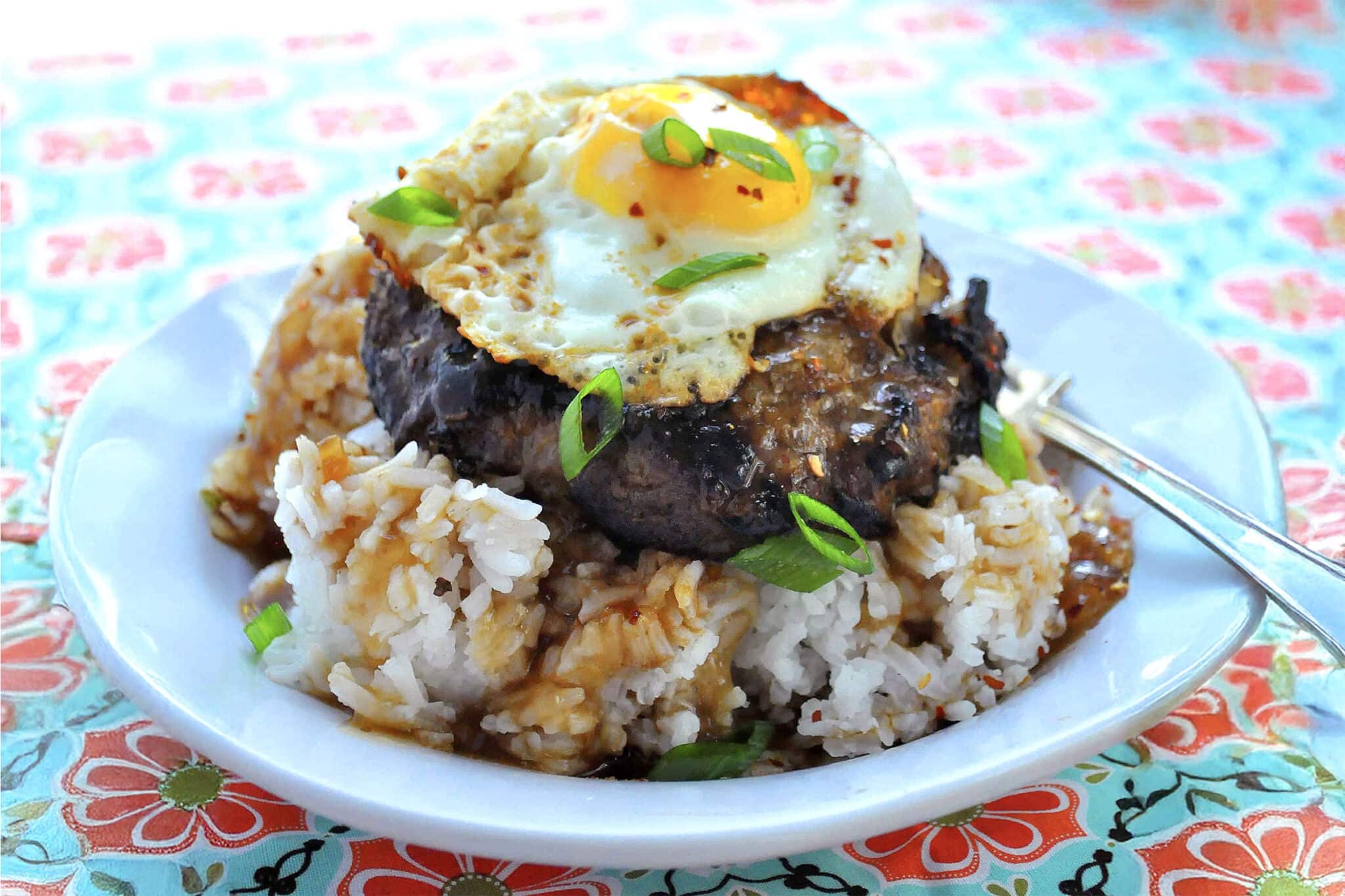 A plate of rice topped with a teriyaki loco moco—juicy hamburger patty, brown gravy, and a sunny-side up egg—finished with chopped green onions on a colorful floral tablecloth.