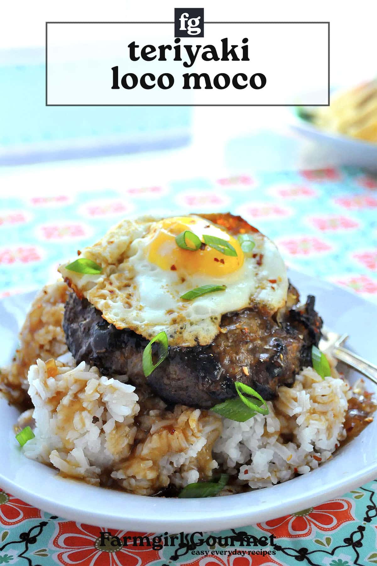 A plate of teriyaki loco moco featuring white rice topped with a hamburger patty, savory brown teriyaki sauce, a fried egg, and chopped green onions rests on a colorful floral tablecloth.