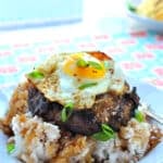 A plate of teriyaki loco moco featuring white rice, a hamburger patty, brown gravy, a sunny-side-up egg, and green onions. Served on a white plate atop a colorful, floral tablecloth.