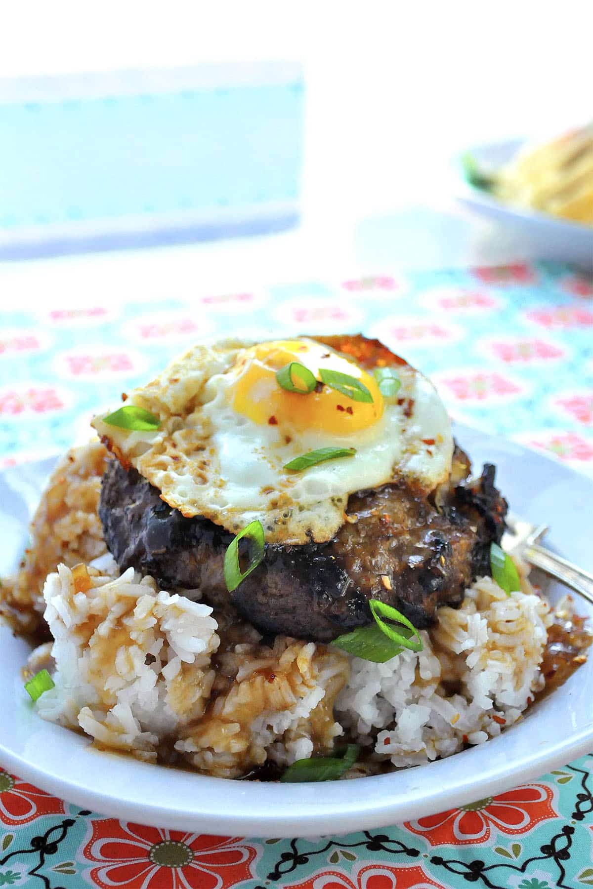 A plate of teriyaki loco moco featuring white rice, a hamburger patty, brown gravy, a sunny-side-up egg, and green onions. Served on a white plate atop a colorful, floral tablecloth.