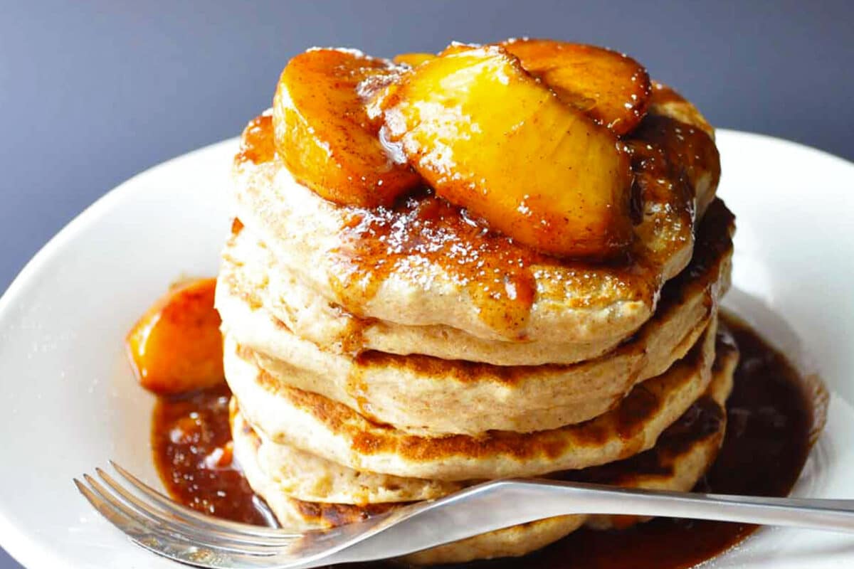 A stack of applesauce pancakes topped with caramelized apple slices and syrup sits on a white plate, with a fork resting in the foreground.