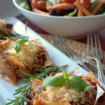 Mini lasagna stacks, reminiscent of easy lasagne muffins, are topped with basil and grated cheese on a white plate, garnished with rosemary. In the background, a mixed green salad with tomatoes and carrots awaits in a large white bowl.