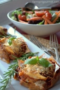Mini lasagna stacks, reminiscent of easy lasagne muffins, are topped with basil and grated cheese on a white plate, garnished with rosemary. In the background, a mixed green salad with tomatoes and carrots awaits in a large white bowl.