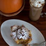 A plate of pumpkin challah bread pudding dusted with powdered sugar, a fork, a glass mug of whipped cream-topped coffee, and a whole pumpkin on a wooden table.
