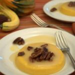 A round serving of pumpkin coconut panna cotta topped with candied pecans and syrup sits on a white plate with a fork. Another plate and decorative gourds are visible in the background on a striped tablecloth.