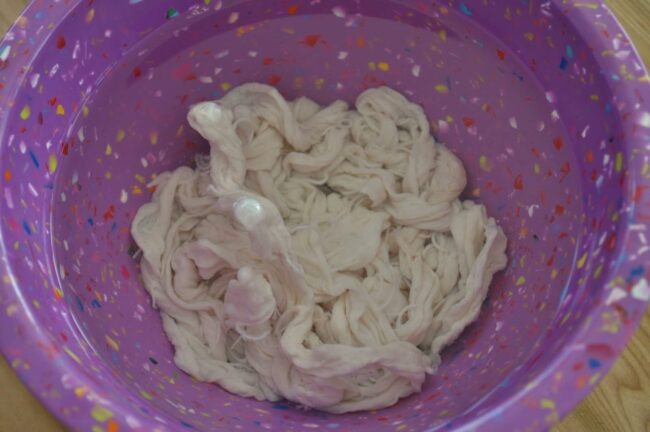 A colorful purple bowl with confetti-like specks contains a white, twisted, and slightly soapy mass of yarn or fabric soaking inside—resembling homemade linguica waiting to be prepared.