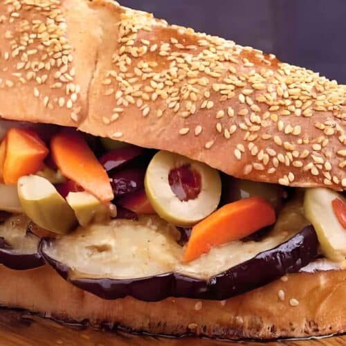 A close-up of an Eggplant Muffuletta sandwich filled with sliced eggplant, olives, carrots, and other pickled vegetables on a sesame seed bun atop a wooden surface.