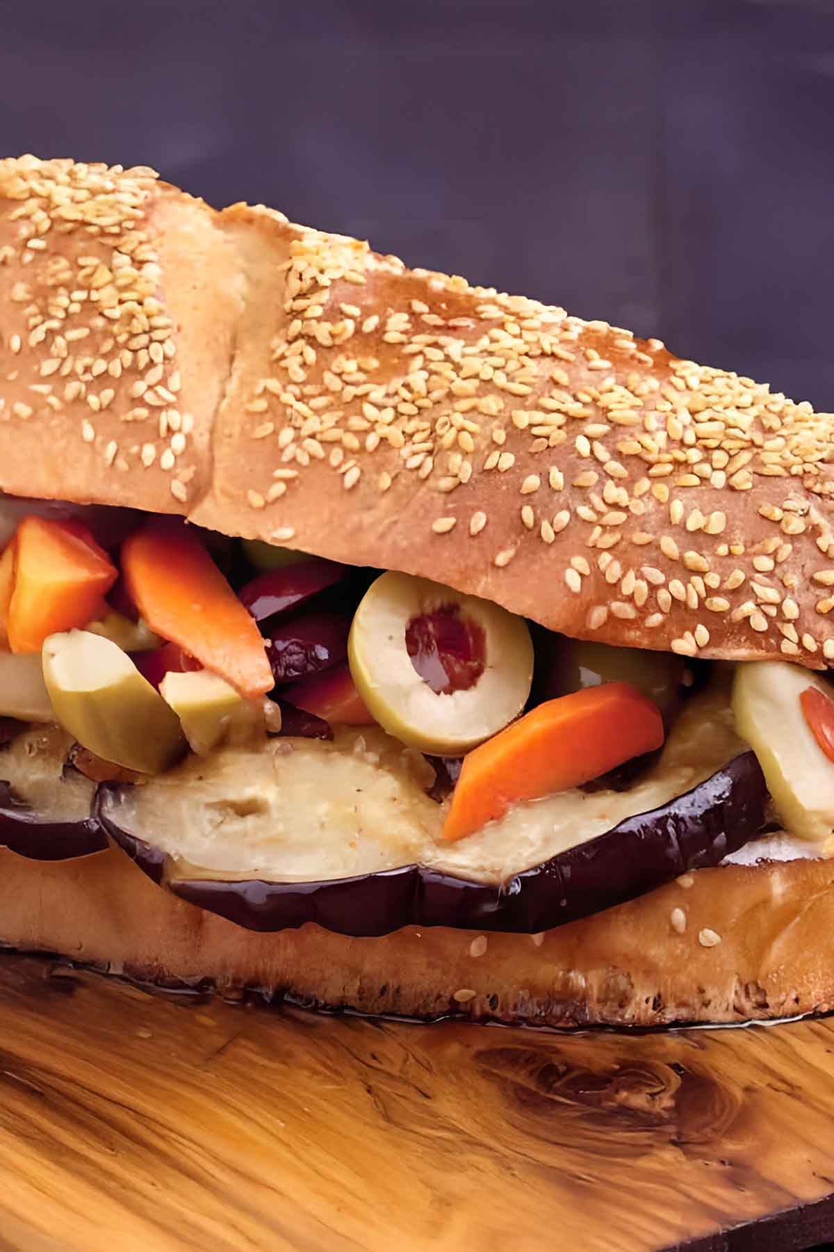 A close-up of an Eggplant Muffuletta sandwich filled with sliced eggplant, olives, carrots, and other pickled vegetables on a sesame seed bun atop a wooden surface.