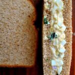 Egg salad with fresh herbs sits atop whole wheat bread, next to a plain slice, ready for sandwich-making. The Farmgirl Gourmet logo is visible in the bottom left corner.