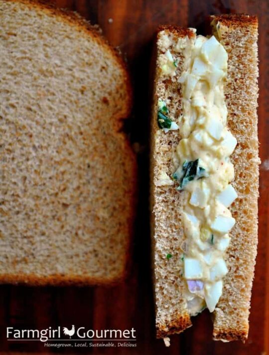 Egg salad with fresh herbs sits atop whole wheat bread, next to a plain slice, ready for sandwich-making. The Farmgirl Gourmet logo is visible in the bottom left corner.