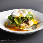 A white plate with a fork holding a bite of a crab cake—reminiscent of savory clam cakes—topped with fresh greens and a poached egg, runny yolk spilling onto the plate. The words Farmgirl Gourmet are visible at the corner.