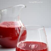 A clear glass and pitcher filled with homemade fresh cranberry juice sit on a white surface. The bright red juice has a frothy layer on top. Text on the image reads “Homemade Fresh Cranberry Juice.”.
