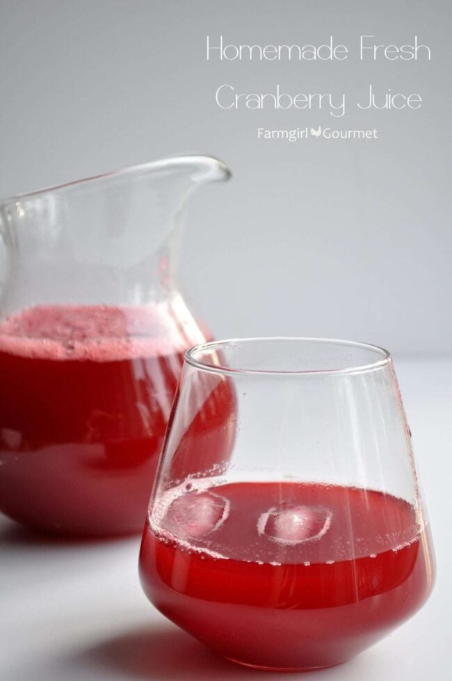A clear glass and pitcher filled with homemade fresh cranberry juice sit on a white surface. The bright red juice has a frothy layer on top. Text on the image reads “Homemade Fresh Cranberry Juice.”.
