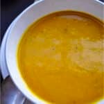 A white bowl filled with creamy orange garam masala soup sits on a matching plate with a spoon resting nearby, set against a dark background.