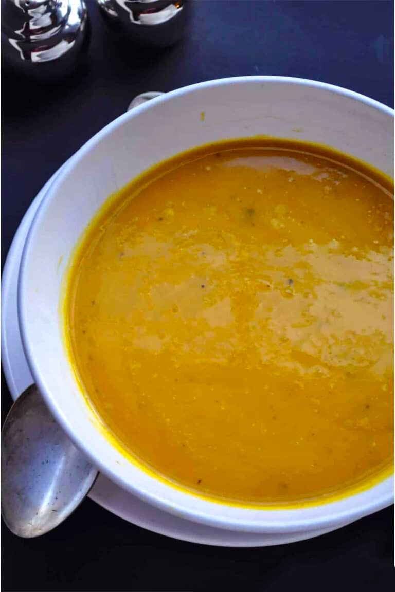 A white bowl filled with creamy orange garam masala soup sits on a matching plate with a spoon resting nearby, set against a dark background.