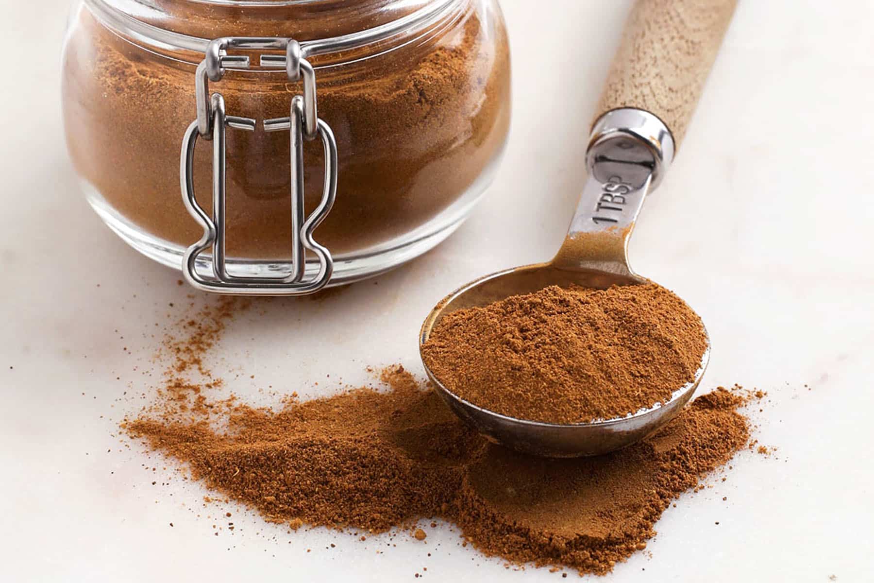 A glass jar of ground pumpkin pie spice next to a wooden-handled measuring spoon filled with the spice.