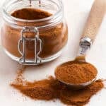A glass jar of pumpkin pie spice next to a measuring spoon filled with the powder, with some spilled on the surface.