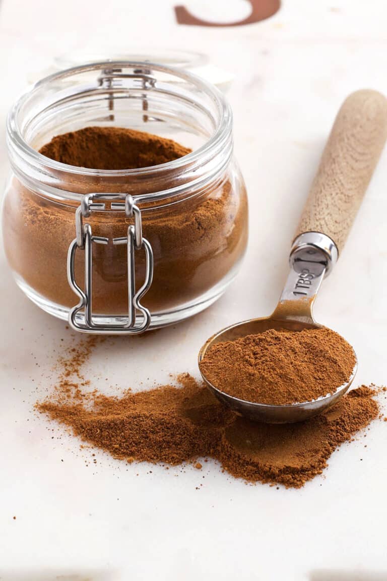 A glass jar of pumpkin pie spice next to a measuring spoon filled with the powder, with some spilled on the surface.