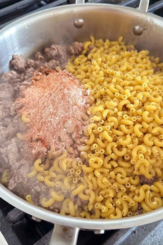 A close-up of a pan on a stove containing cooked ground beef, dry elbow macaroni, and a pile of seasoning mix, ready to be stirred together for a homemade hamburger helper one-pot meal. Steam is rising from the pan.
