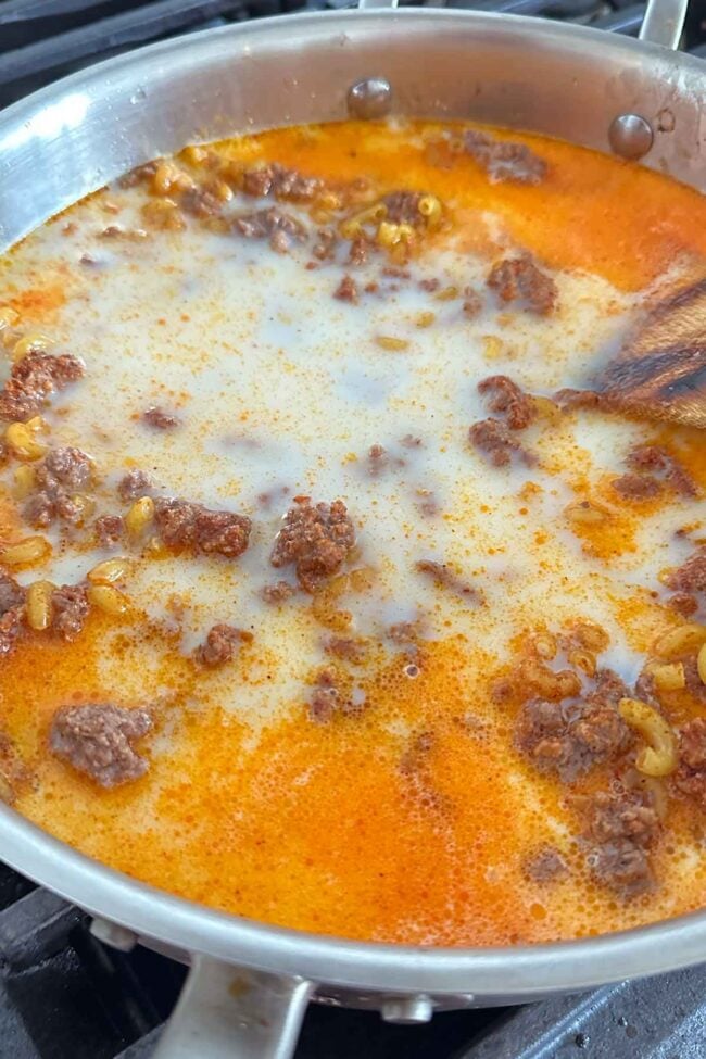 Ground beef, macaroni pasta, and a creamy orange sauce simmer in a large metal skillet on the stovetop—a wooden spoon stirring this homemade hamburger helper to perfection.