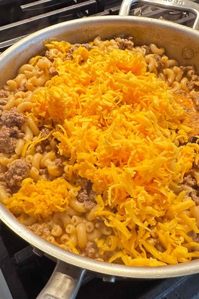 A skillet on a stove filled with cooked macaroni, ground beef, and a large pile of shredded cheddar cheese on top - this homemade hamburger helper is ready to be mixed together.