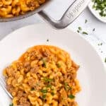 A plate of creamy macaroni with ground meat in a rich orange sauce, garnished with chopped parsley—just like a homemade hamburger helper. A pot with more of this one pot hamburger helper and fresh parsley are nearby, with a fork resting on the plate.