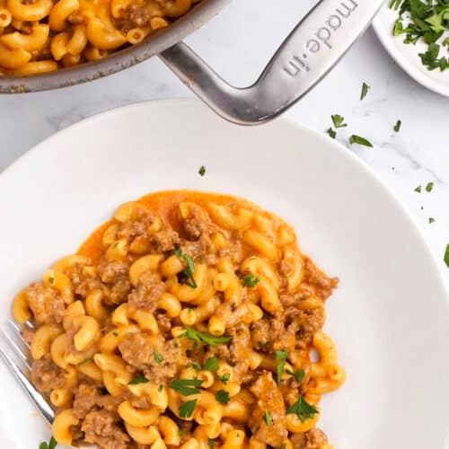 A plate of creamy macaroni with ground meat in a rich orange sauce, garnished with chopped parsley—just like a homemade hamburger helper. A pot with more of this one pot hamburger helper and fresh parsley are nearby, with a fork resting on the plate.