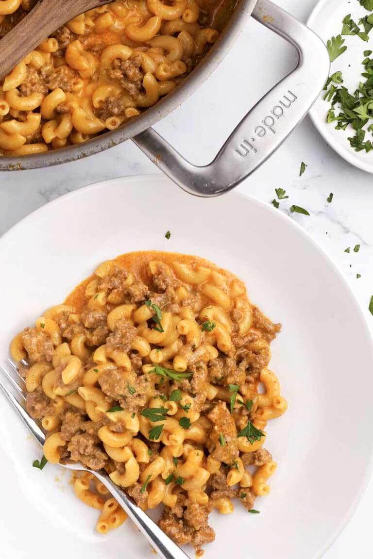 A plate of creamy macaroni with ground meat in a rich orange sauce, garnished with chopped parsley—just like a homemade hamburger helper. A pot with more of this one pot hamburger helper and fresh parsley are nearby, with a fork resting on the plate.