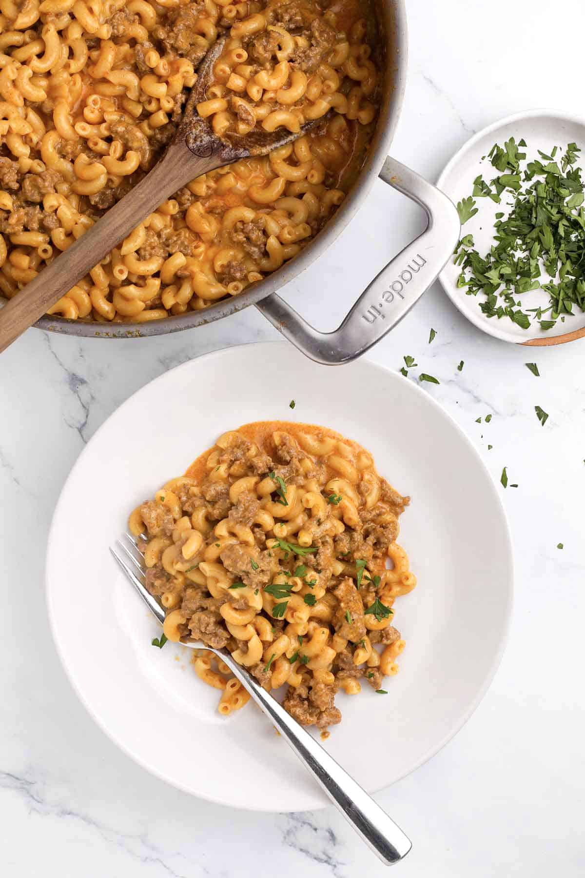 A white plate with cheesy macaroni and ground beef—paired with hamburger helper seasoning mix—is garnished with chopped parsley beside a pan of the same easy weeknight dinner and a bowl of extra parsley. A fork rests on the plate.