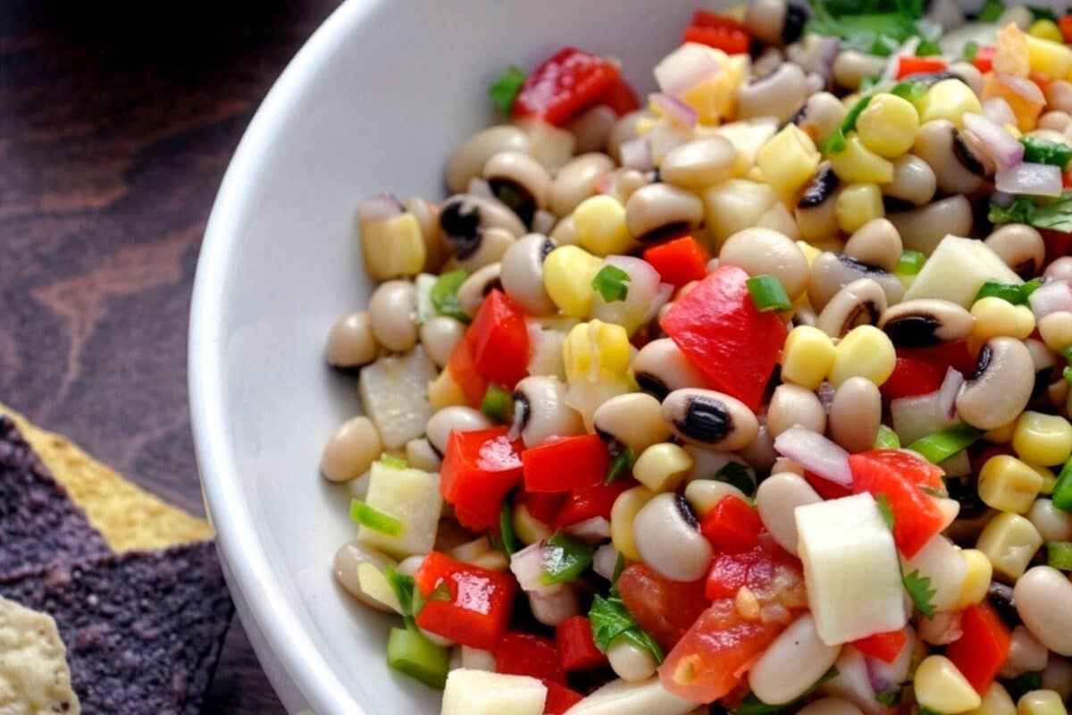 A close-up of a bowl filled with colorful Texas caviar, featuring diced red peppers, corn, onions, green herbs, and black-eyed peas, with tortilla chips visible on the side.