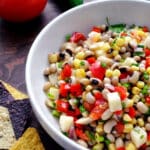 A white bowl filled with colorful Texas caviar, featuring black-eyed peas, corn, tomatoes, onions, and peppers. Blue and yellow tortilla chips sit beside the bowl. A tomato and a jalapeño are in the background.