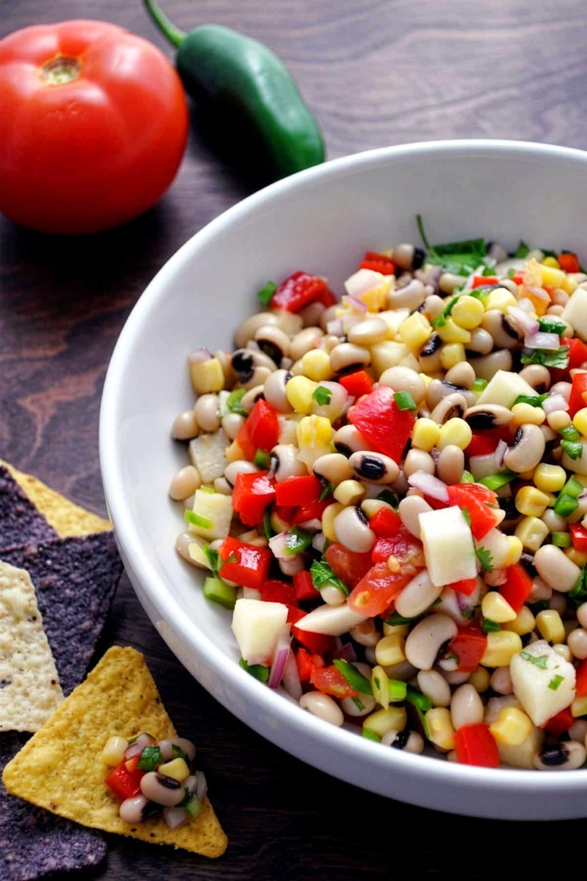 A white bowl filled with colorful Texas caviar, featuring black-eyed peas, corn, tomatoes, onions, and peppers. Blue and yellow tortilla chips sit beside the bowl. A tomato and a jalapeño are in the background.