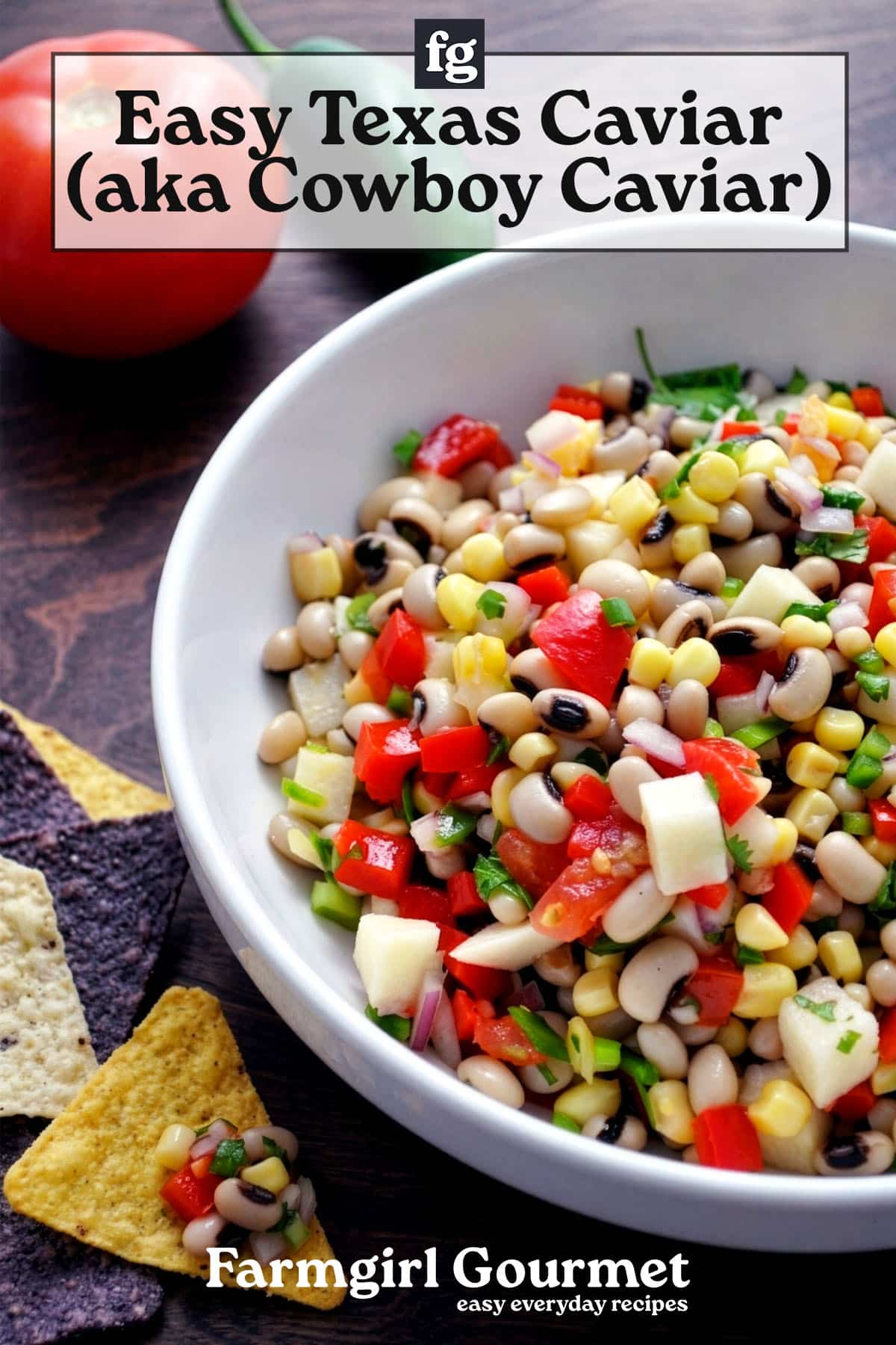 A bowl of Texas caviar salad with black-eyed peas, corn, diced tomatoes, onions, and peppers sits on a table with tortilla chips and a tomato in the background. Text overlay reads Easy Texas Caviar recipe (aka Cowboy Caviar).