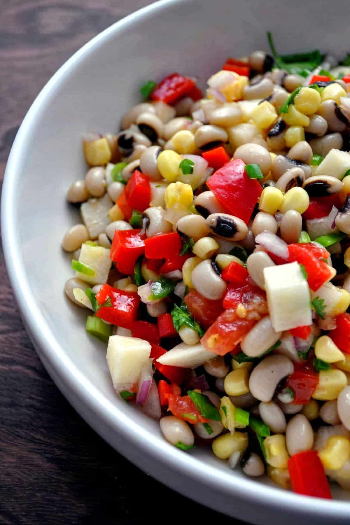 A close-up of a bowl filled with vibrant texas caviar—a colorful salad of black-eyed peas, diced red peppers, corn, green onions, and cubes of white cheese or vegetables—mixed together and garnished with fresh herbs.