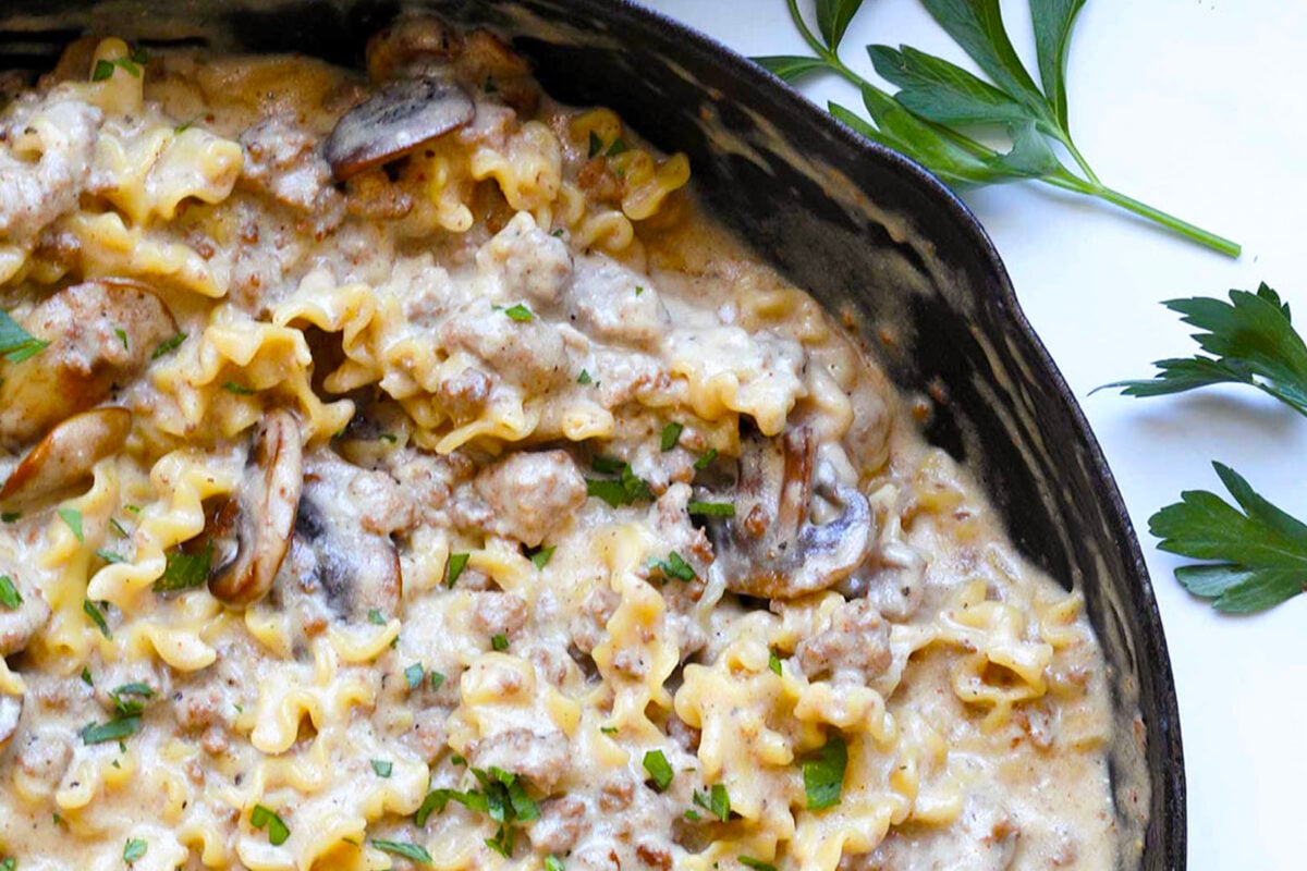 A skillet filled with creamy beef stroganoff hamburger helper, ground meat, mushrooms, and chopped parsley, with fresh parsley leaves scattered on a white surface beside the pan.