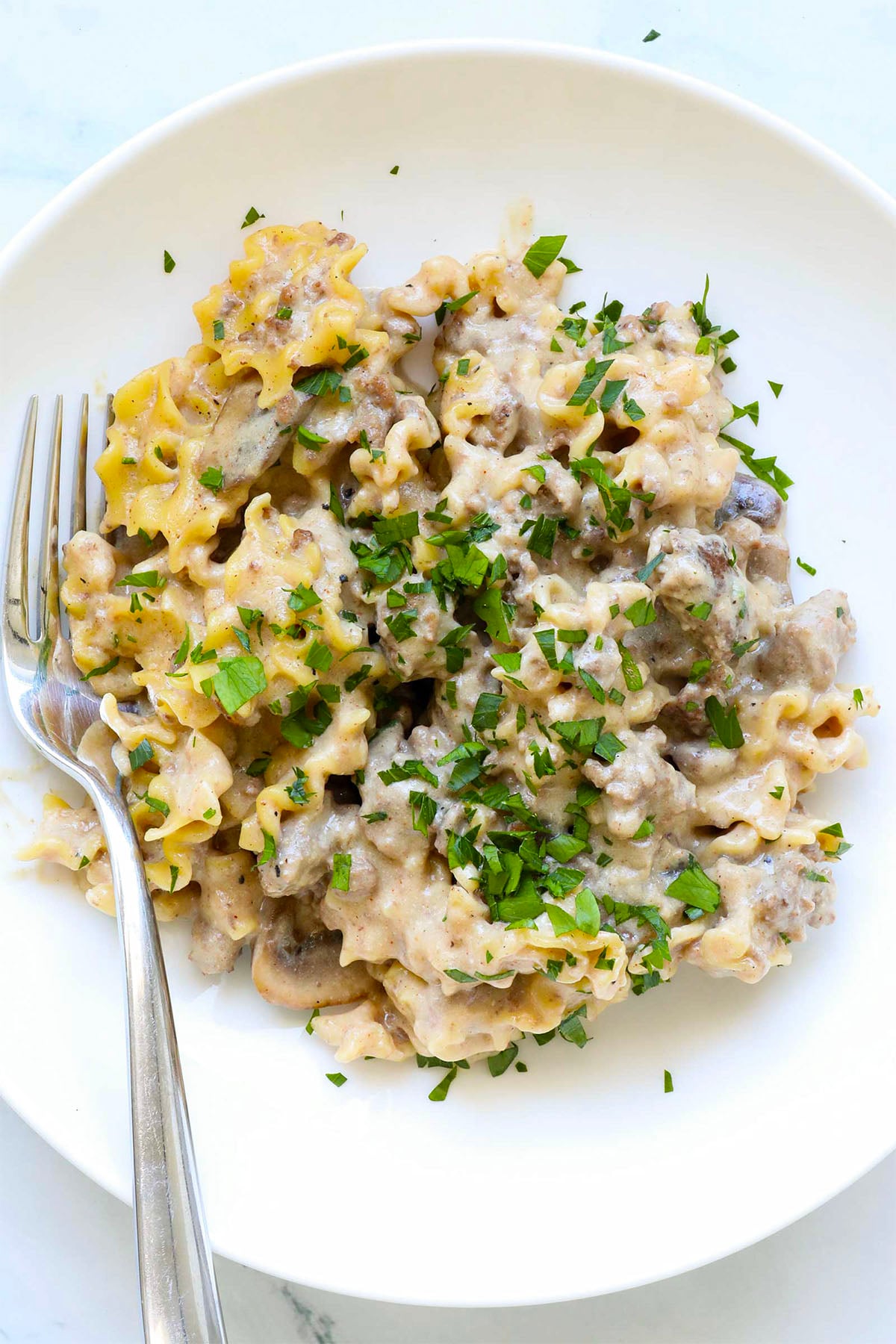 A white plate with creamy beef stroganoff hamburger helper pasta, topped with chopped fresh parsley, sits invitingly next to a fork on the left side of the plate.