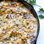A cast-iron skillet filled with creamy beef stroganoff hamburger helper, mixed with ground meat and mushrooms, is garnished with chopped parsley and sits on a white surface beside fresh parsley sprigs.
