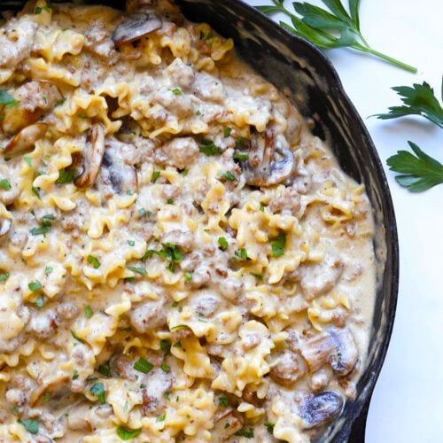 A cast-iron skillet filled with creamy beef stroganoff hamburger helper, mixed with ground meat and mushrooms, is garnished with chopped parsley and sits on a white surface beside fresh parsley sprigs.