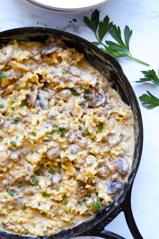 A cast-iron skillet filled with creamy beef stroganoff hamburger helper, mixed with ground meat and mushrooms, is garnished with chopped parsley and sits on a white surface beside fresh parsley sprigs.