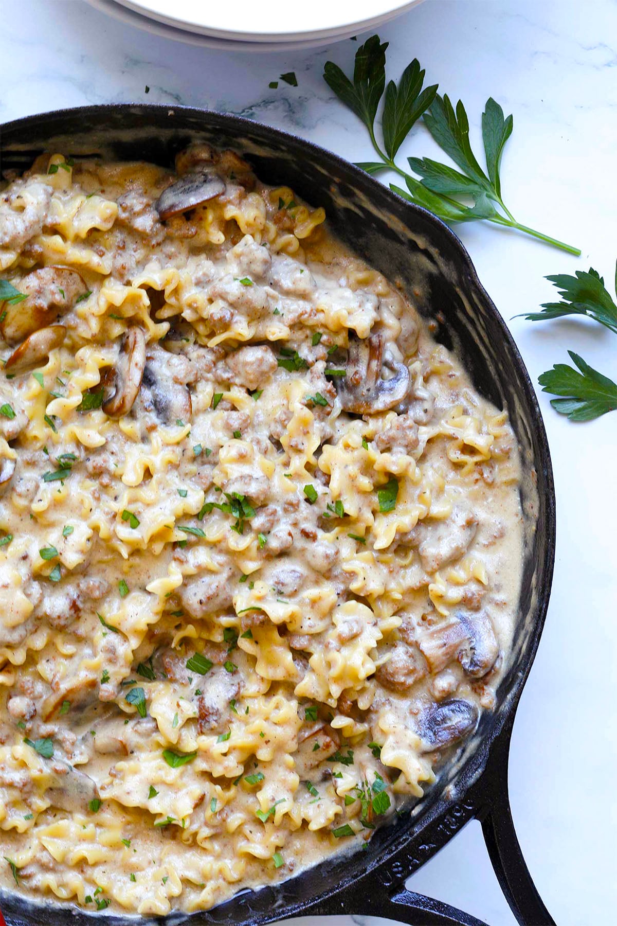 A cast-iron skillet filled with creamy beef stroganoff hamburger helper, mixed with ground meat and mushrooms, is garnished with chopped parsley and sits on a white surface beside fresh parsley sprigs.