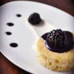 A small brown butter cake topped with blackberry sauce and a fresh blackberry, served on a white plate with cream and blackberry sauce dots for decoration.