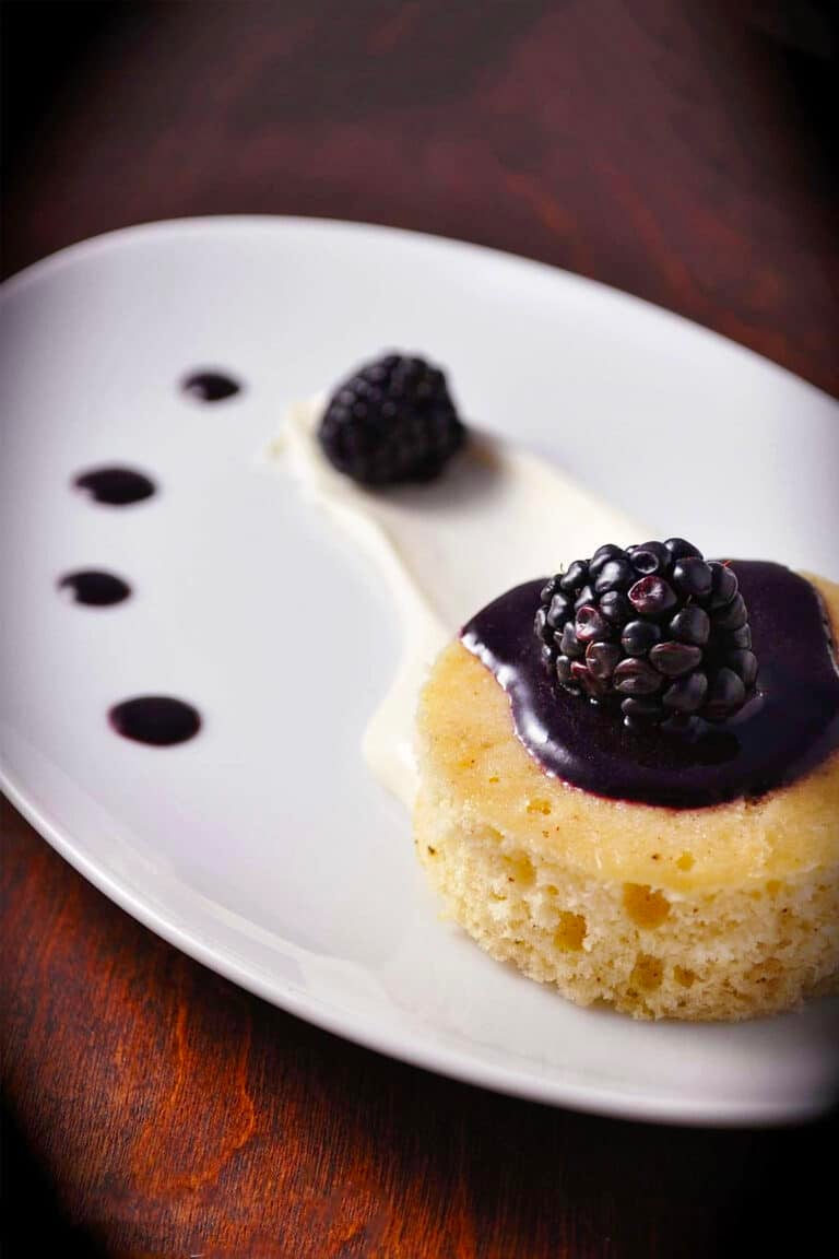 A small brown butter cake topped with blackberry sauce and a fresh blackberry, served on a white plate with cream and blackberry sauce dots for decoration.