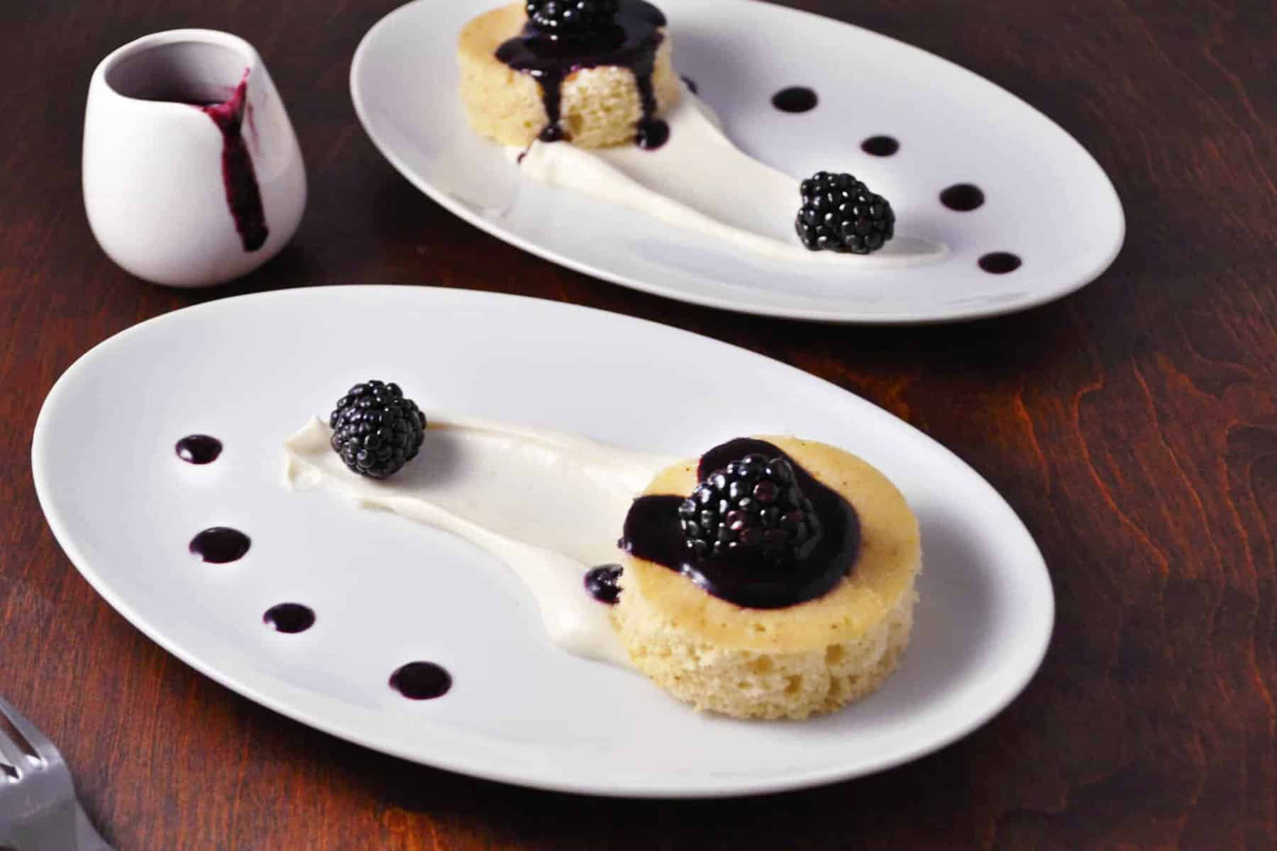 Two white plates with round brown butter cakes, cream, blackberry sauce, and fresh blackberries. A small white pitcher with more sauce is beside the plates on a dark wooden table.