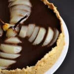 A dark chocolate pear tart with a crumbly crust, topped with sliced poached pears in a fan shape and covered in glossy ganache, is displayed on a white plate against a dark background.